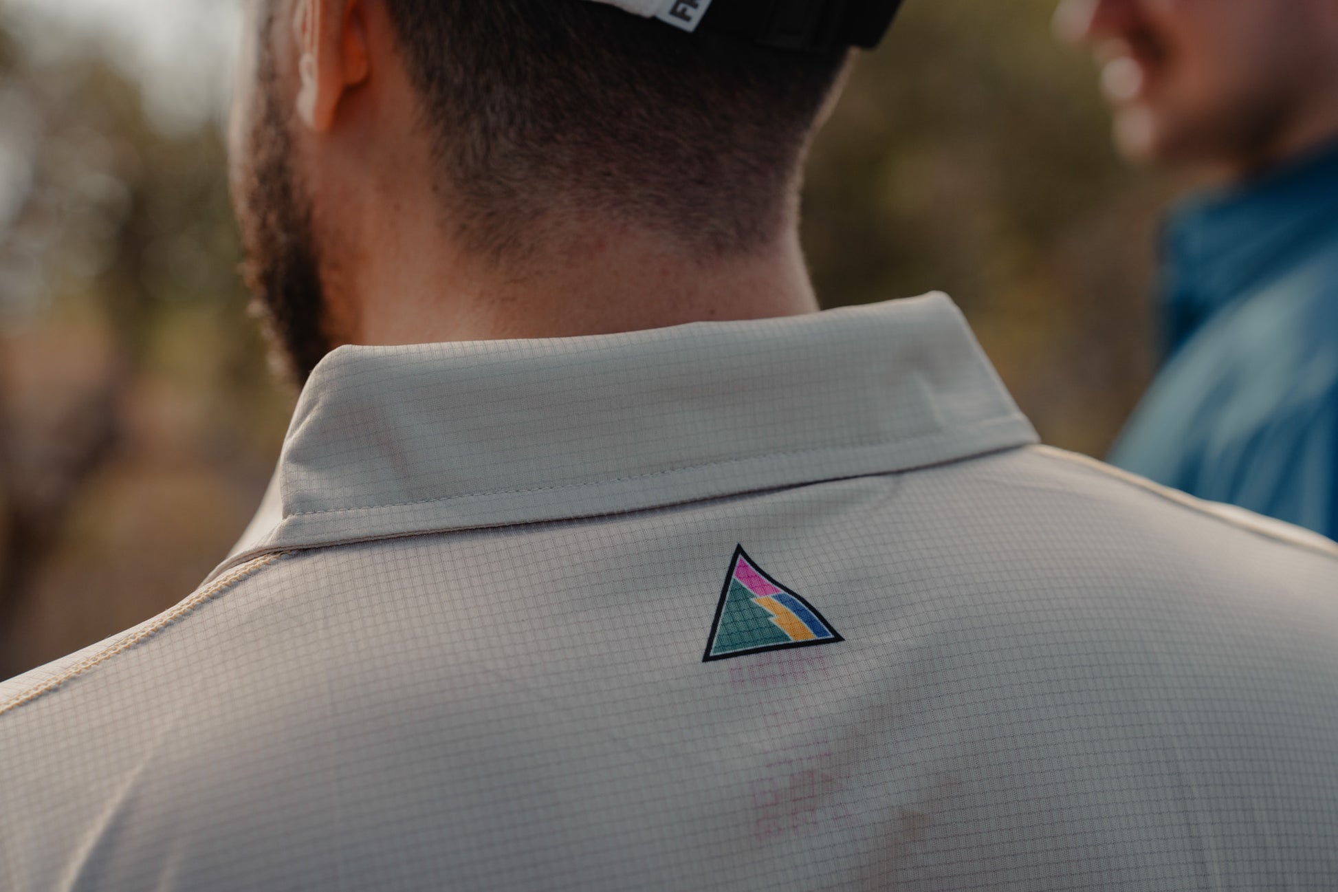 Person wearing a light gray shirt with a colorful triangle logo on the back, blurred background